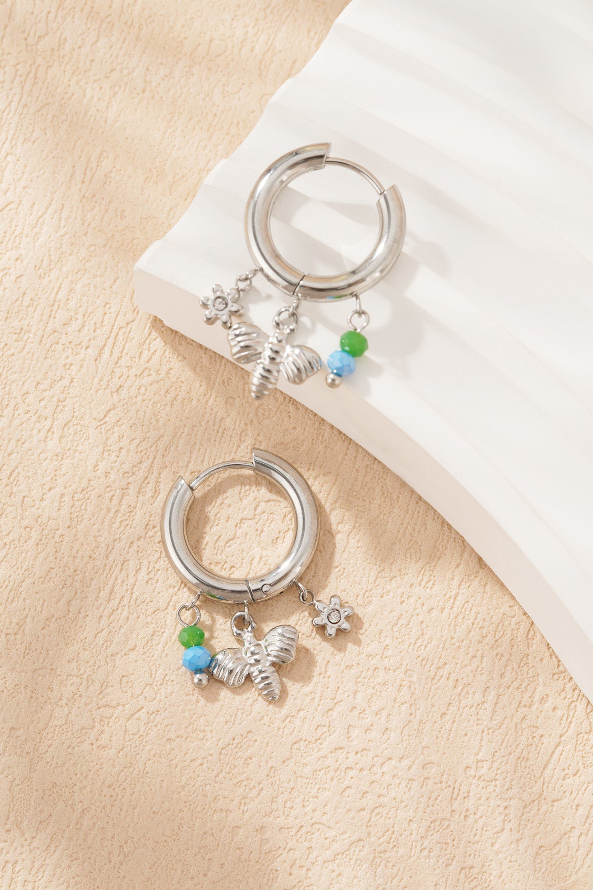 Silver charm hoop earrings with bee, flower charms and green-blue beads on white sculptured surface