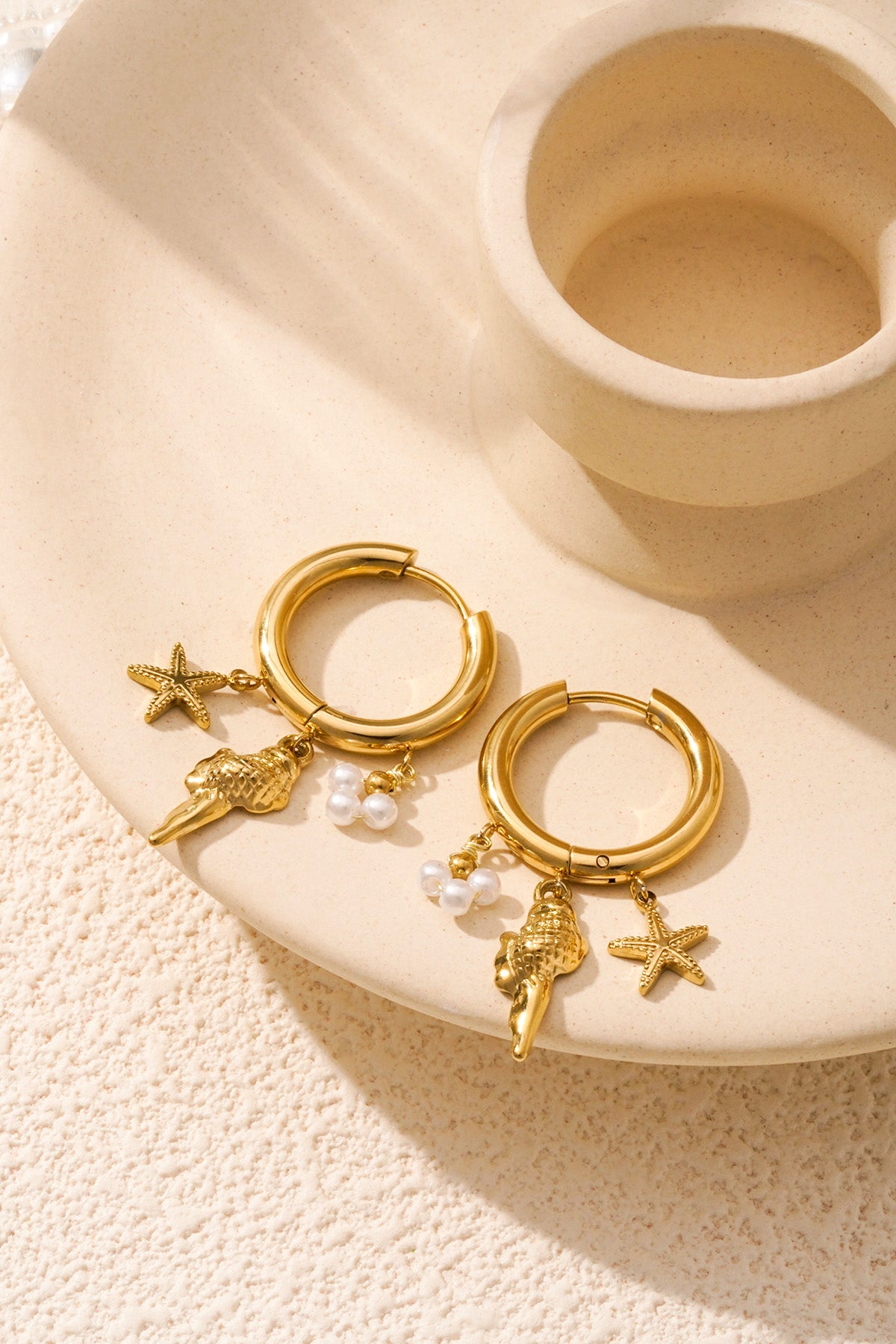 Gold hoop earrings with starfish, shell, and pearl charms styled on beige textured surface