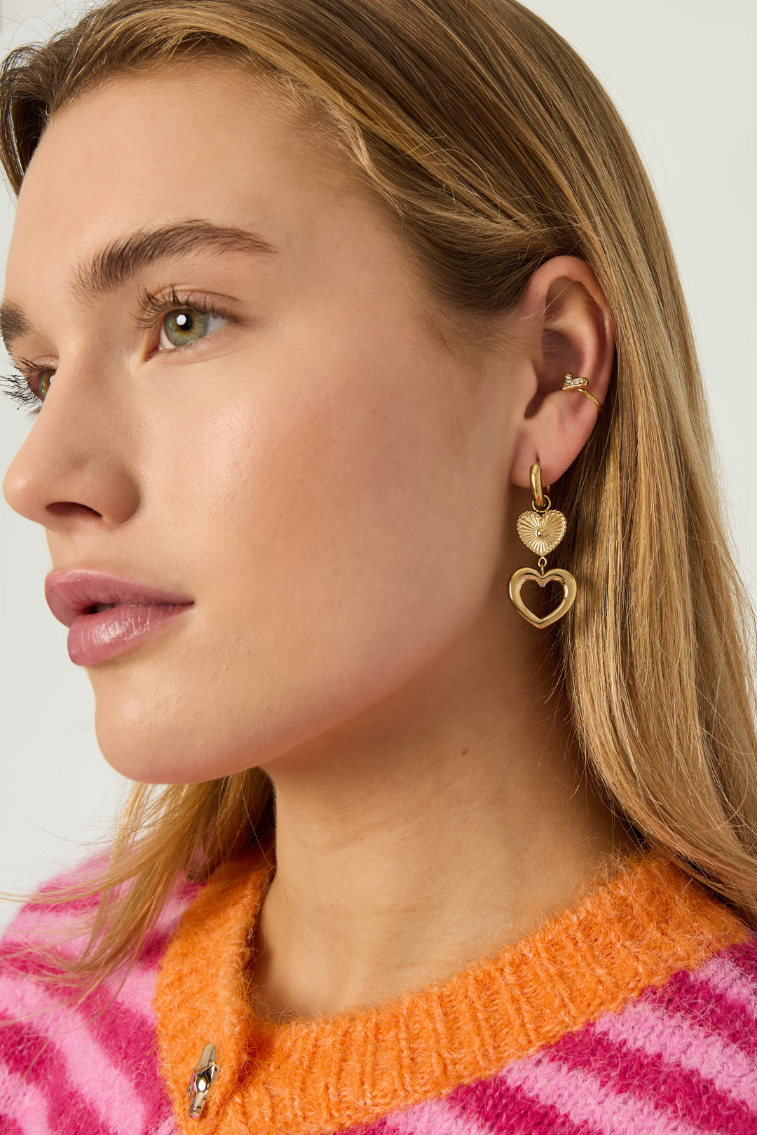 Close-up of a woman wearing gold heart charm hoop earrings paired with a colorful pink and orange sweater
