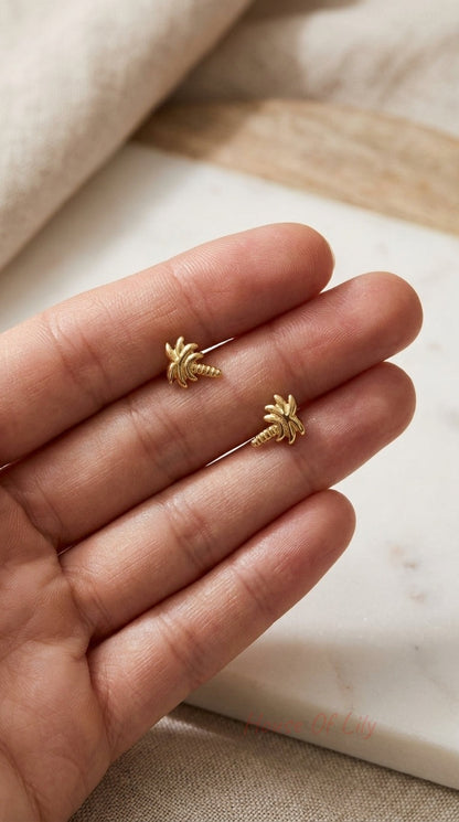 Dainty gold palm tree stud earrings with textured detail, held between fingers against a marble and linen background.
