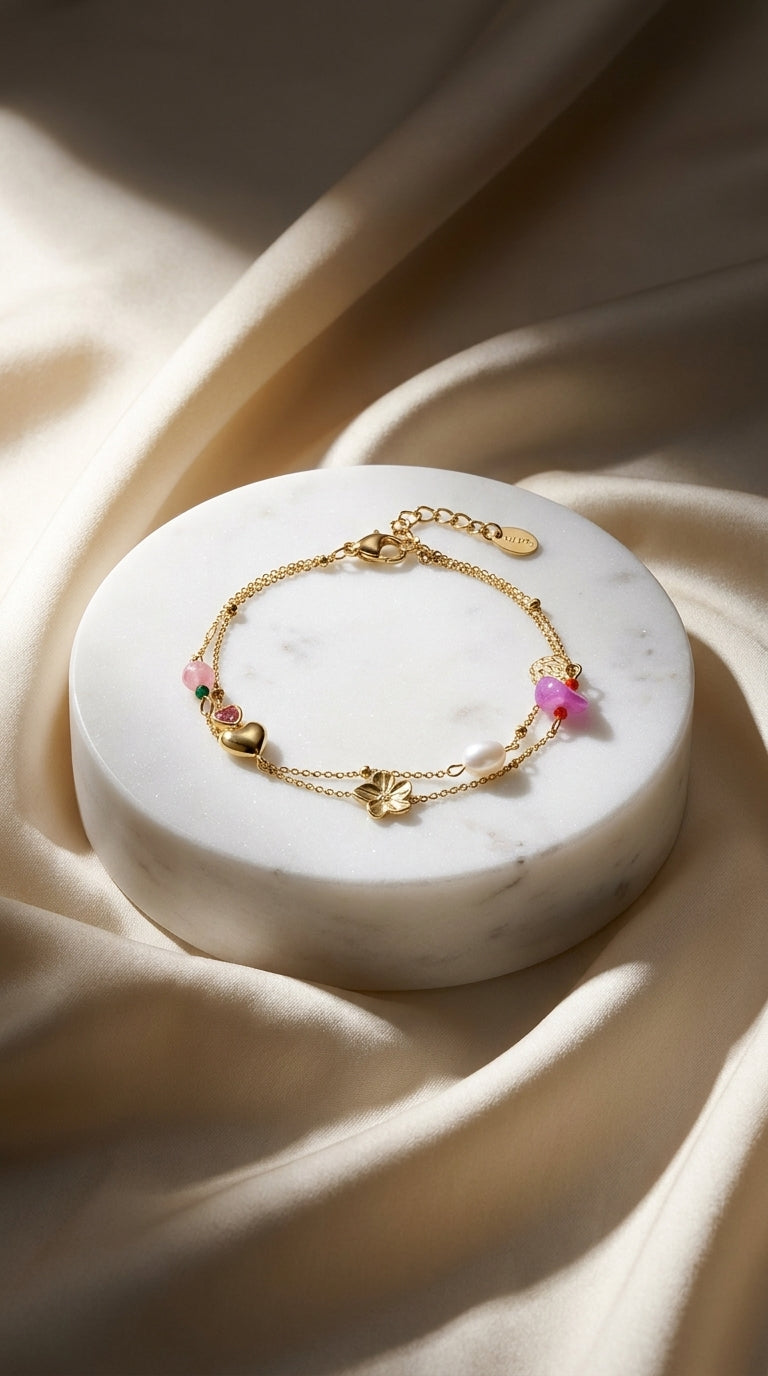 Gold double-strand charm bracelet featuring a flower, heart, pearl, and colorful pink and green stone accents.