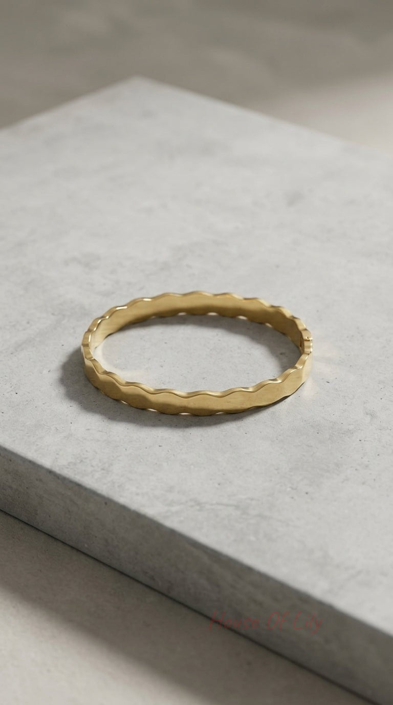 Minimalist gold bangle featuring a delicate, wavy scalloped edge, resting on a textured grey stone surface.