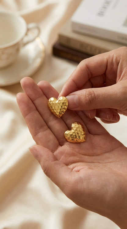 Gold-toned heart earrings with a textured, hammered surface finish held in a palm.