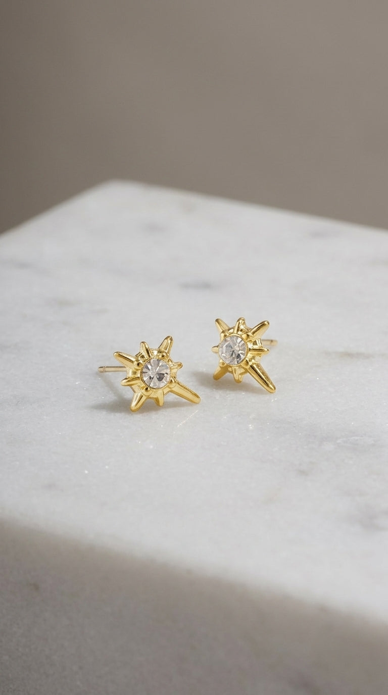 Gold starburst stud earrings featuring a central sparkling crystal on a white marble surface.