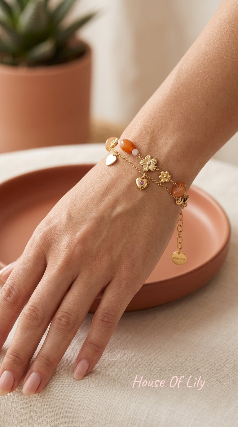 Gold charm bracelet stack with orange gemstones, flower charms, and a heart pendant with a small pink stone.