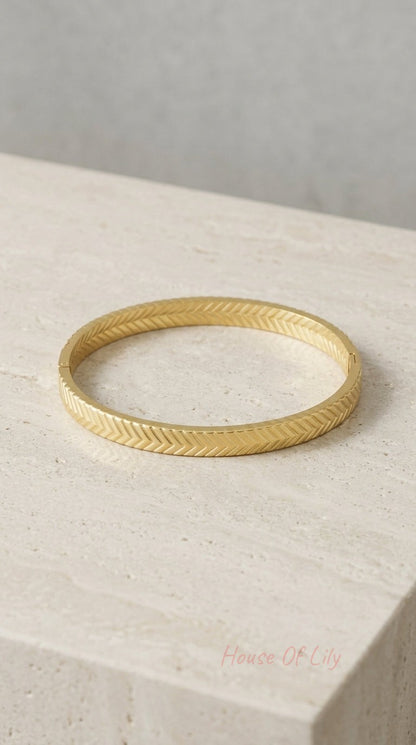 Gold-tone hinged bangle bracelet with a textured herringbone chevron pattern, resting on a beige stone surface.