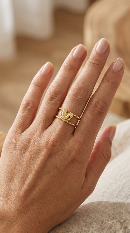 Gold Double Trouble ring on a finger, featuring two beaded bands joined by a central smooth puffed heart charm.