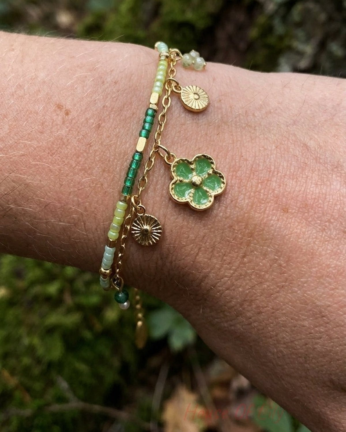Two-strand gold bracelet with green seed beads and dangling flower charms on a wrist.