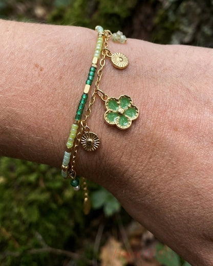 Two-strand gold bracelet with green seed beads and dangling flower charms on a wrist.