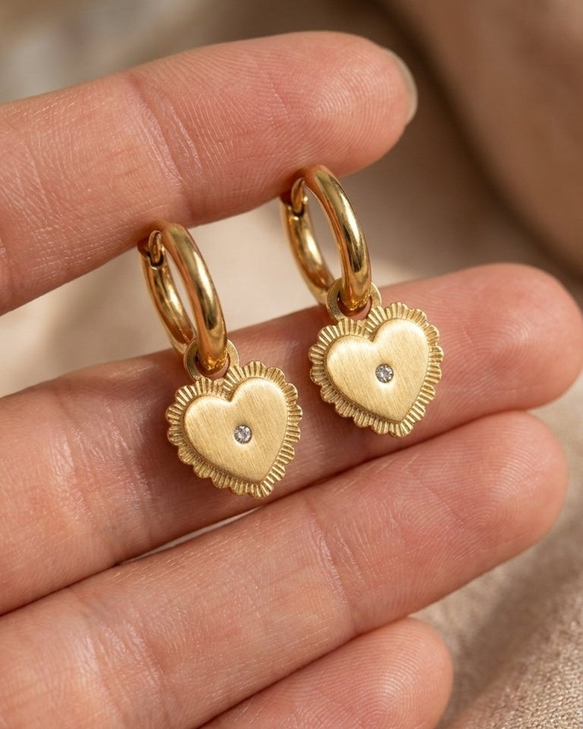 Gold drop earrings featuring puffy, textured heart charms with a small center stone, held in a hand.
