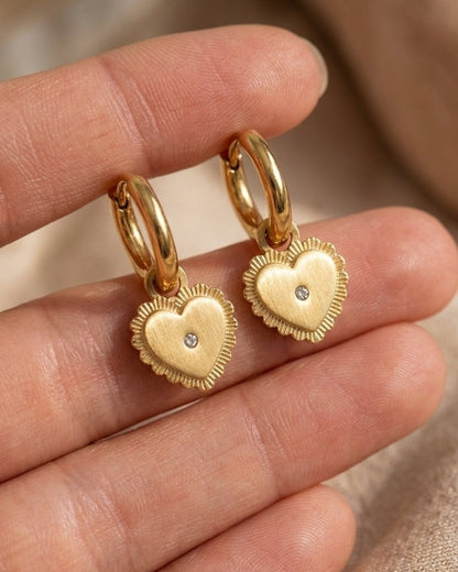 Gold drop earrings featuring puffy, textured heart charms with a small center stone, held in a hand.