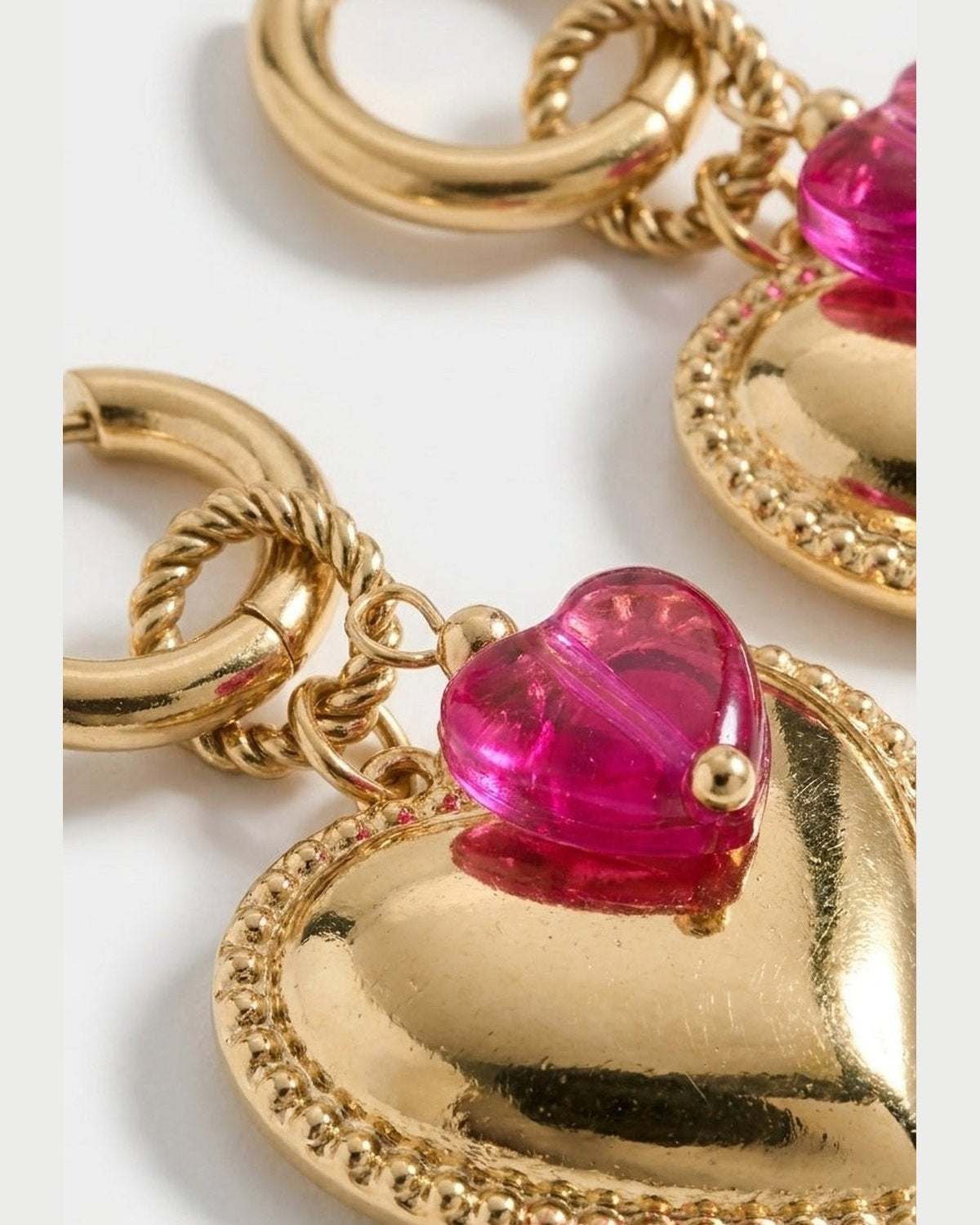 Gold hoop earrings featuring a large puffed heart pendant and a smaller translucent pink crystal heart.