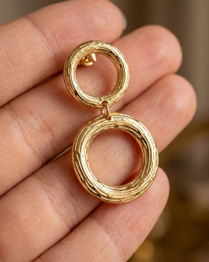 Chic gold circle earrings with a textured, brushed pattern, featuring two linked circular drops held on a hand.