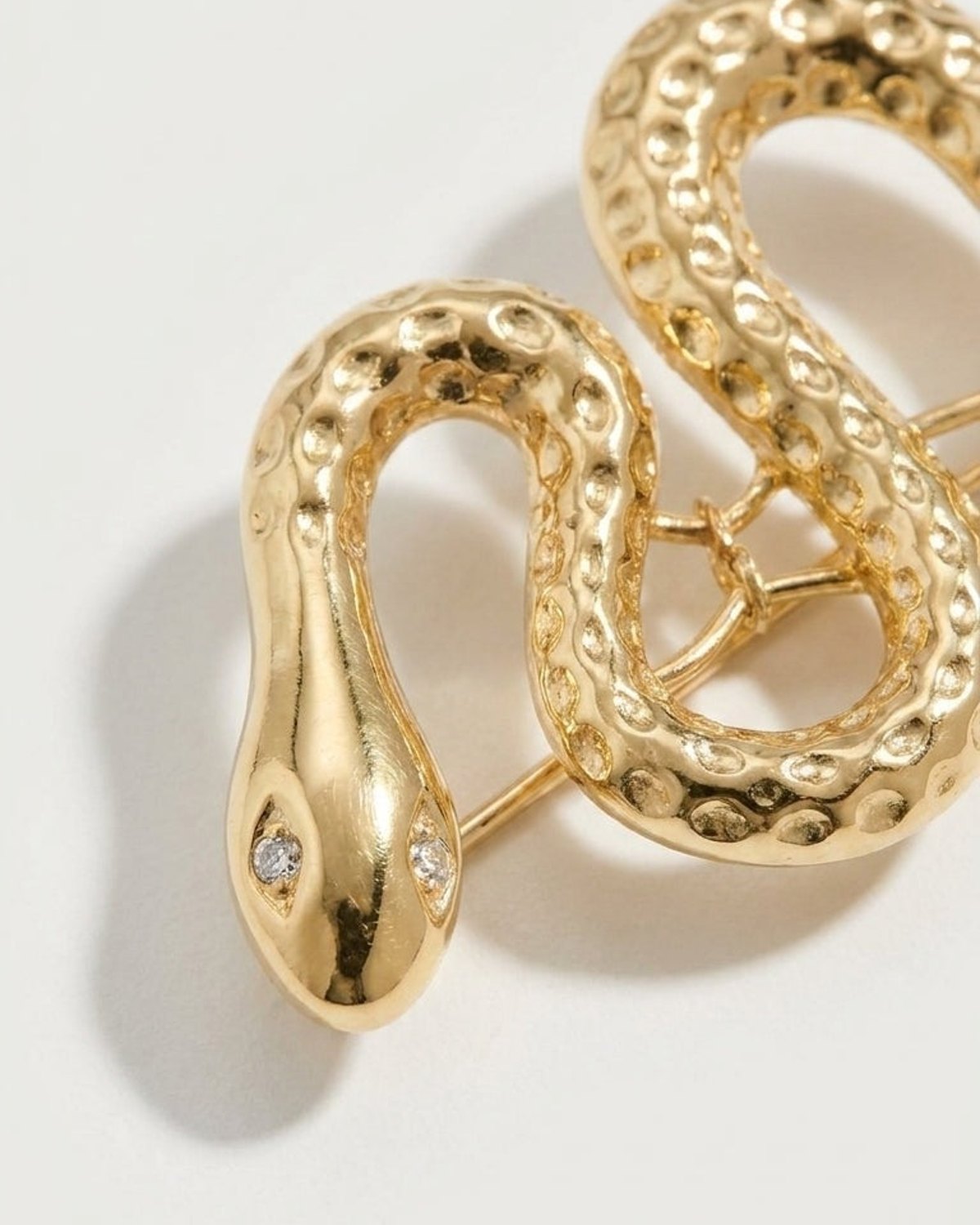 Gold-toned snake brooch featuring textured scales and crystal eyes on a light background.