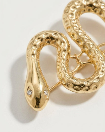 Gold-toned snake brooch featuring textured scales and crystal eyes on a light background.
