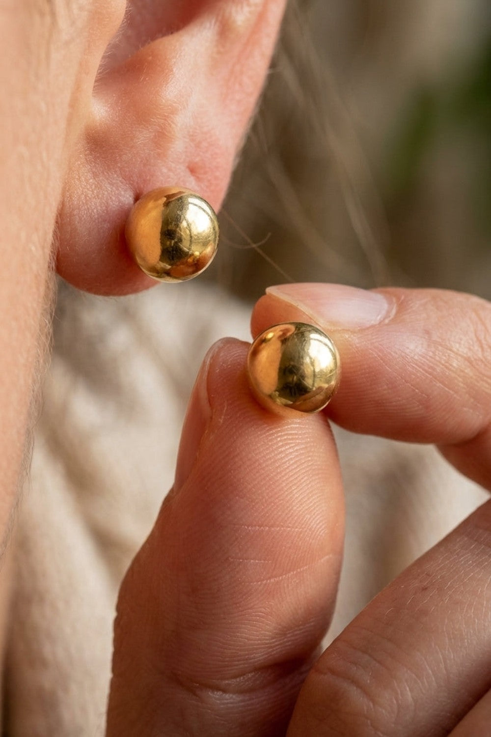 Chic Steel Geometric Studs: polished gold-colored spherical ball stud earrings, one worn on an earlobe and one held in fingers.