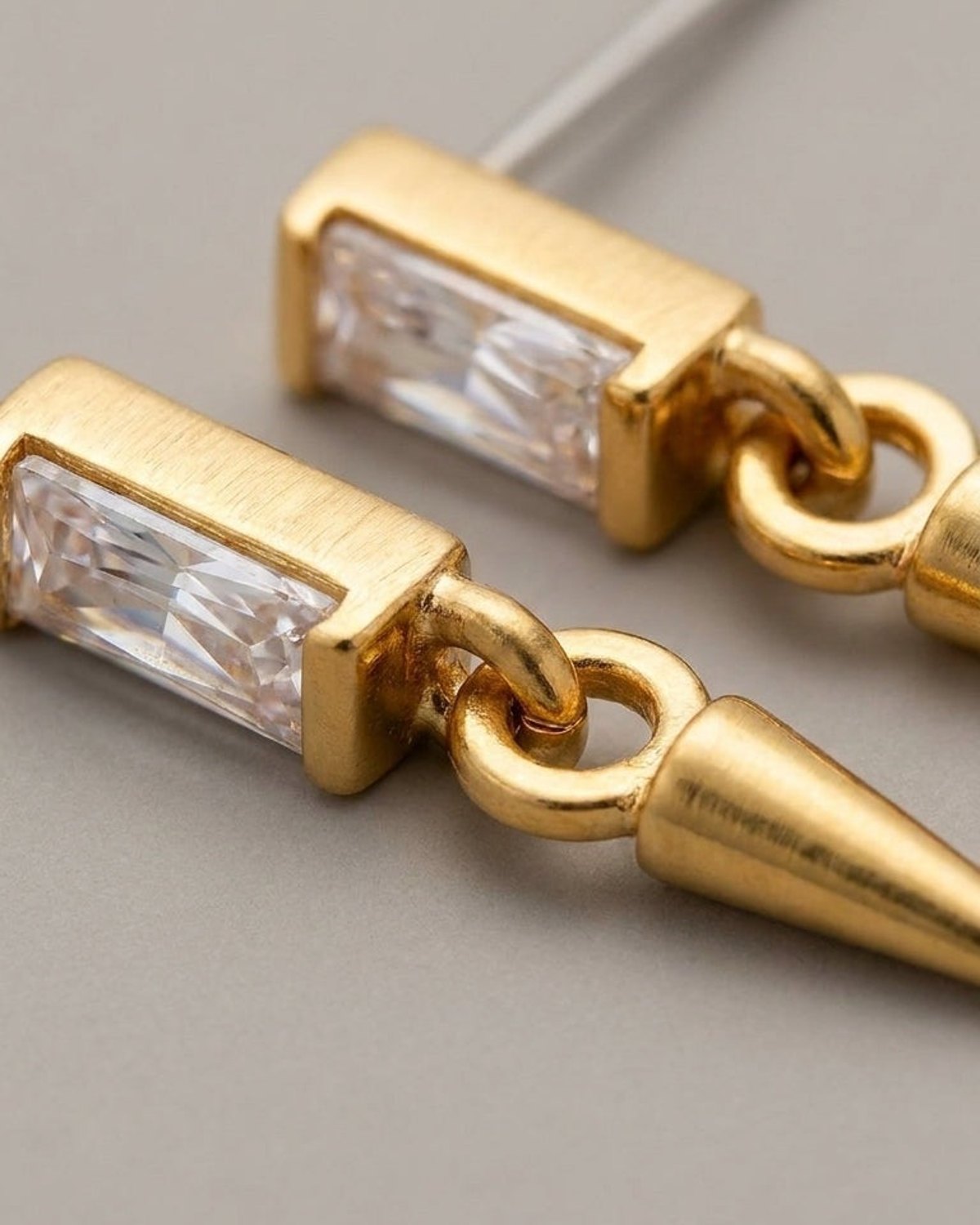 Close-up of gold-toned drop earrings featuring a rectangular clear crystal stud and a hanging cone pendant.