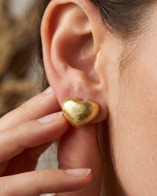A hand holding a textured gold heart stud earring worn on an earlobe.