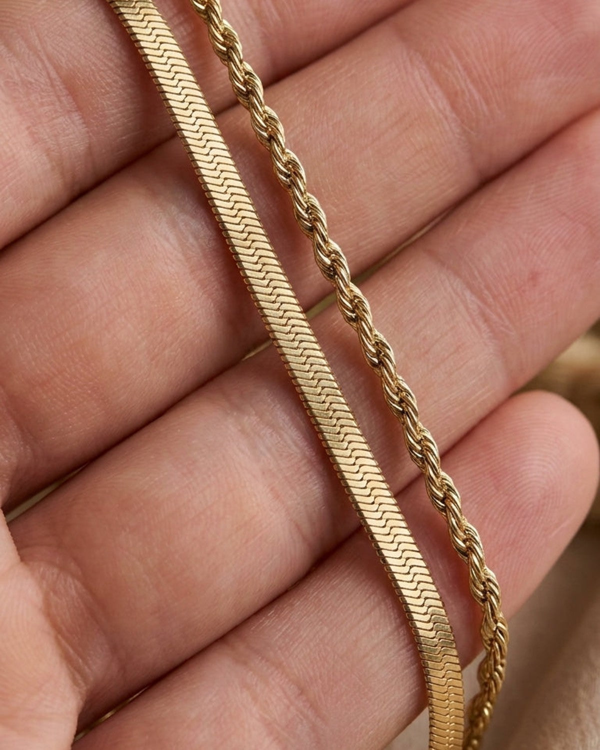 Gold dual chain necklace featuring a flat herringbone chain and a twisted rope chain held in a hand.