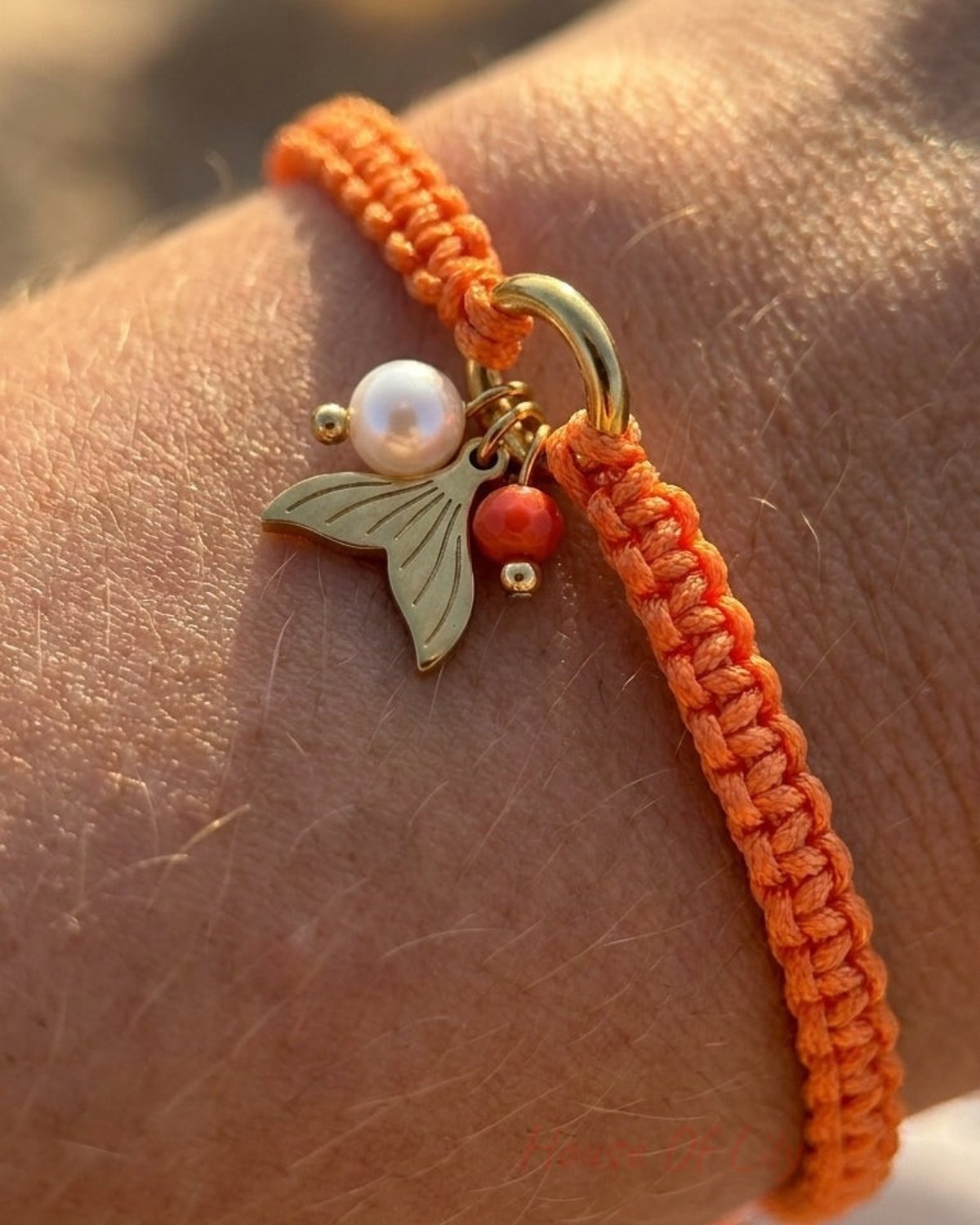 Orange braided cord bracelet with a gold ring and mermaid tail, pearl, and coral charms on a wrist.