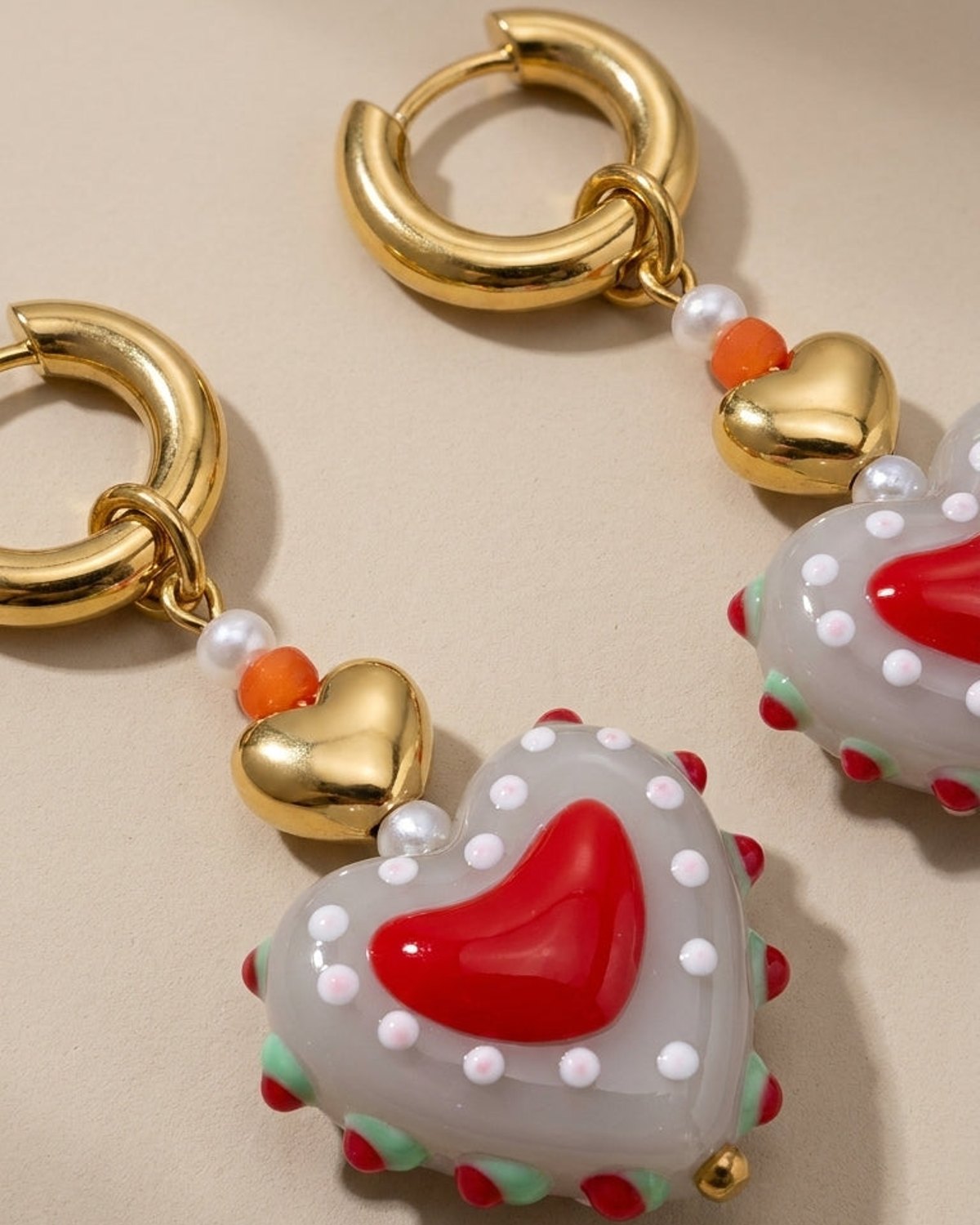 Gold hoop earrings with charms of small beads, a gold heart, and a large grey heart with a red center and dots.