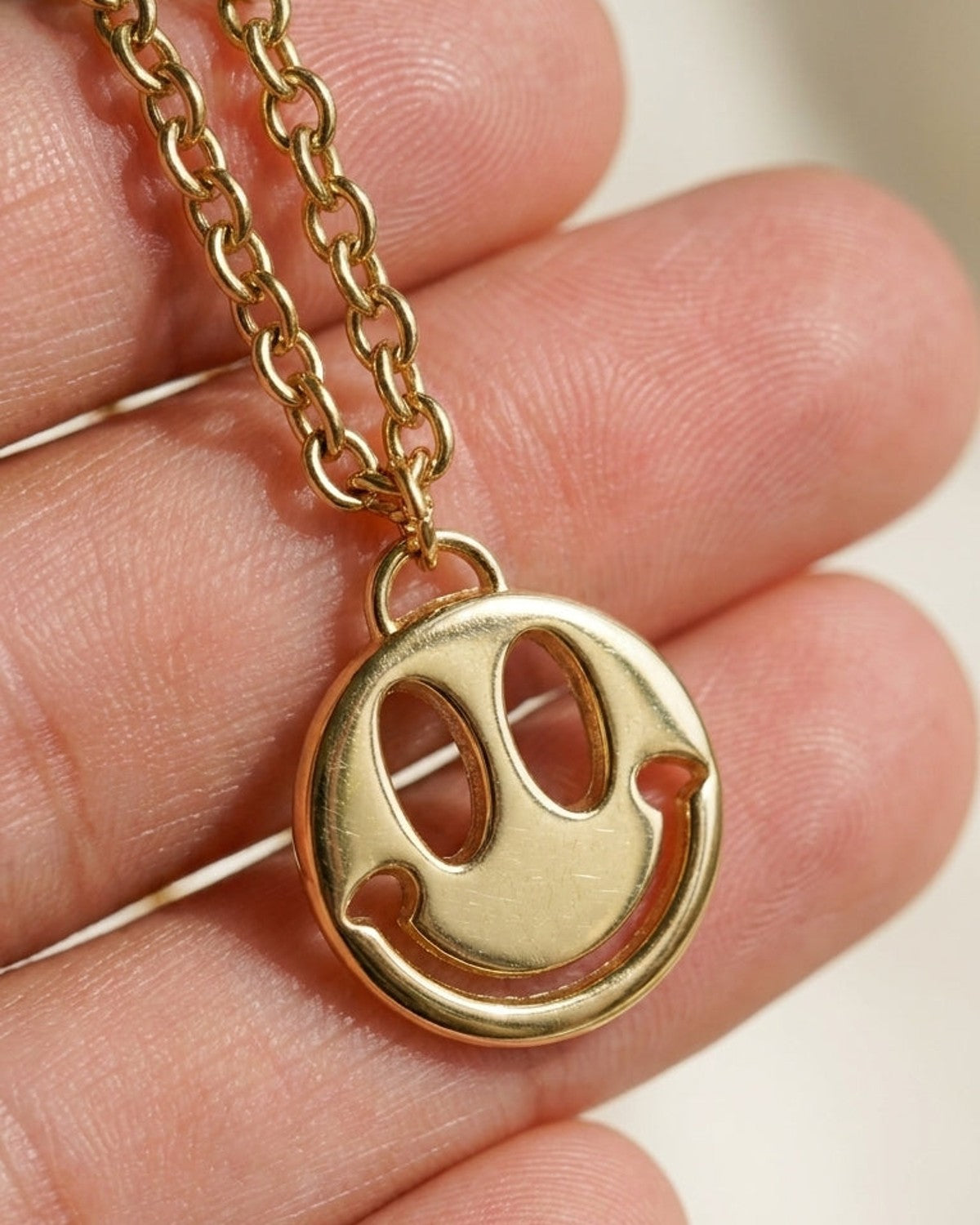 Gold chain necklace featuring a round smiley face pendant resting on a person's fingers.