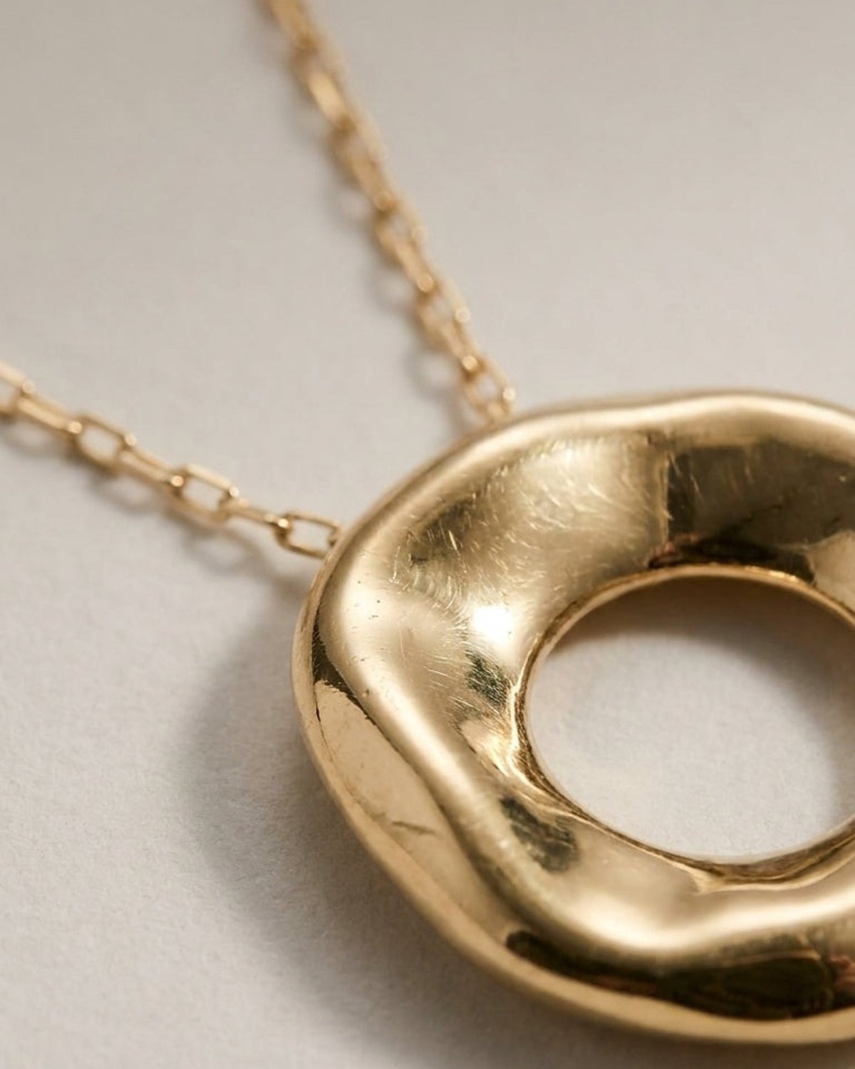 A gold necklace with a thick, irregularly shaped circular pendant on a matching chain against a neutral background.