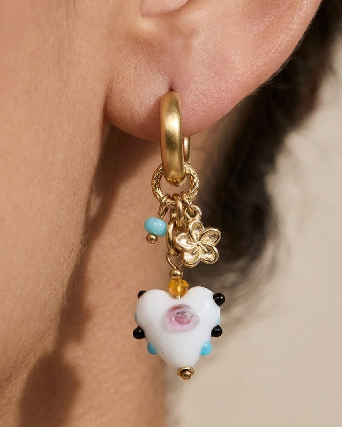 Gold huggie hoop earring with dangling flower, blue bead, and a white glass heart with pink and black details.