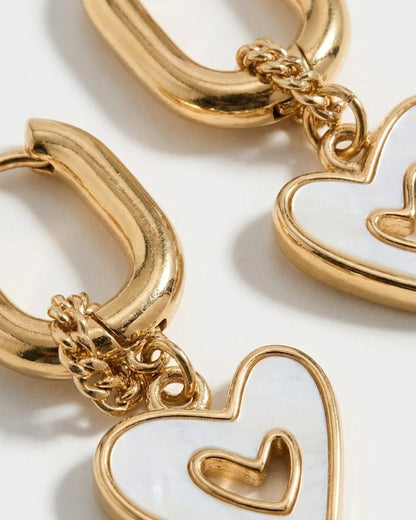 Gold drop earrings with thick oval huggie hoops and dangling mother-of-pearl heart charms featuring a cutout heart.