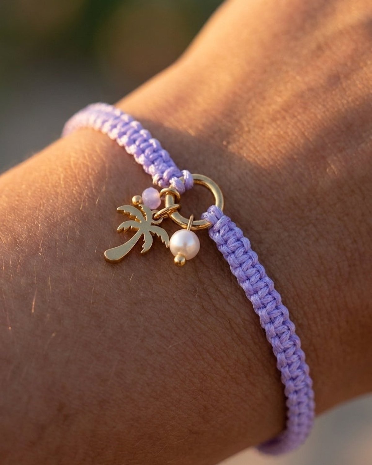 Lilac braided cord bracelet with gold palm tree and pearl charms worn on a wrist.