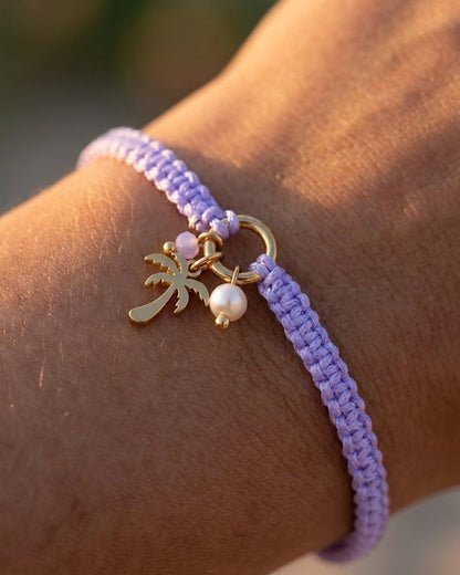 Lilac braided cord bracelet with gold palm tree and pearl charms worn on a wrist.