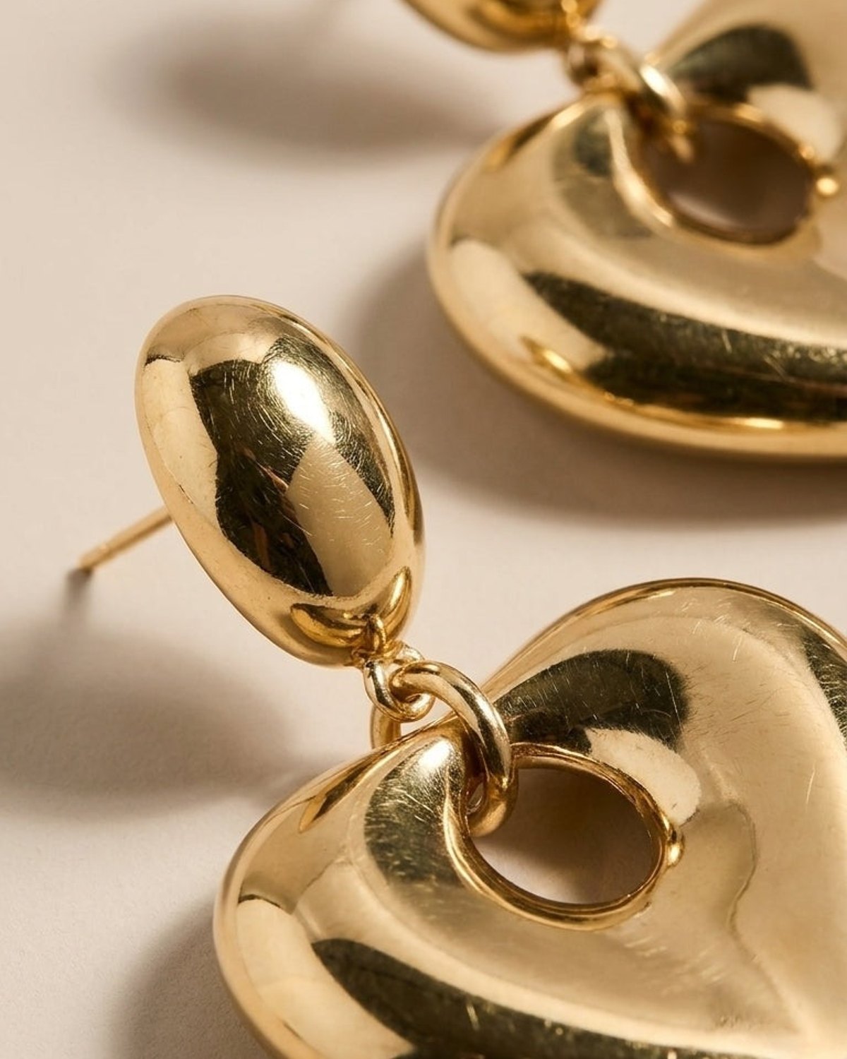 Glossy gold-tone earrings with an oval stud and a dangling, puffed heart pendant featuring a circular cutout.