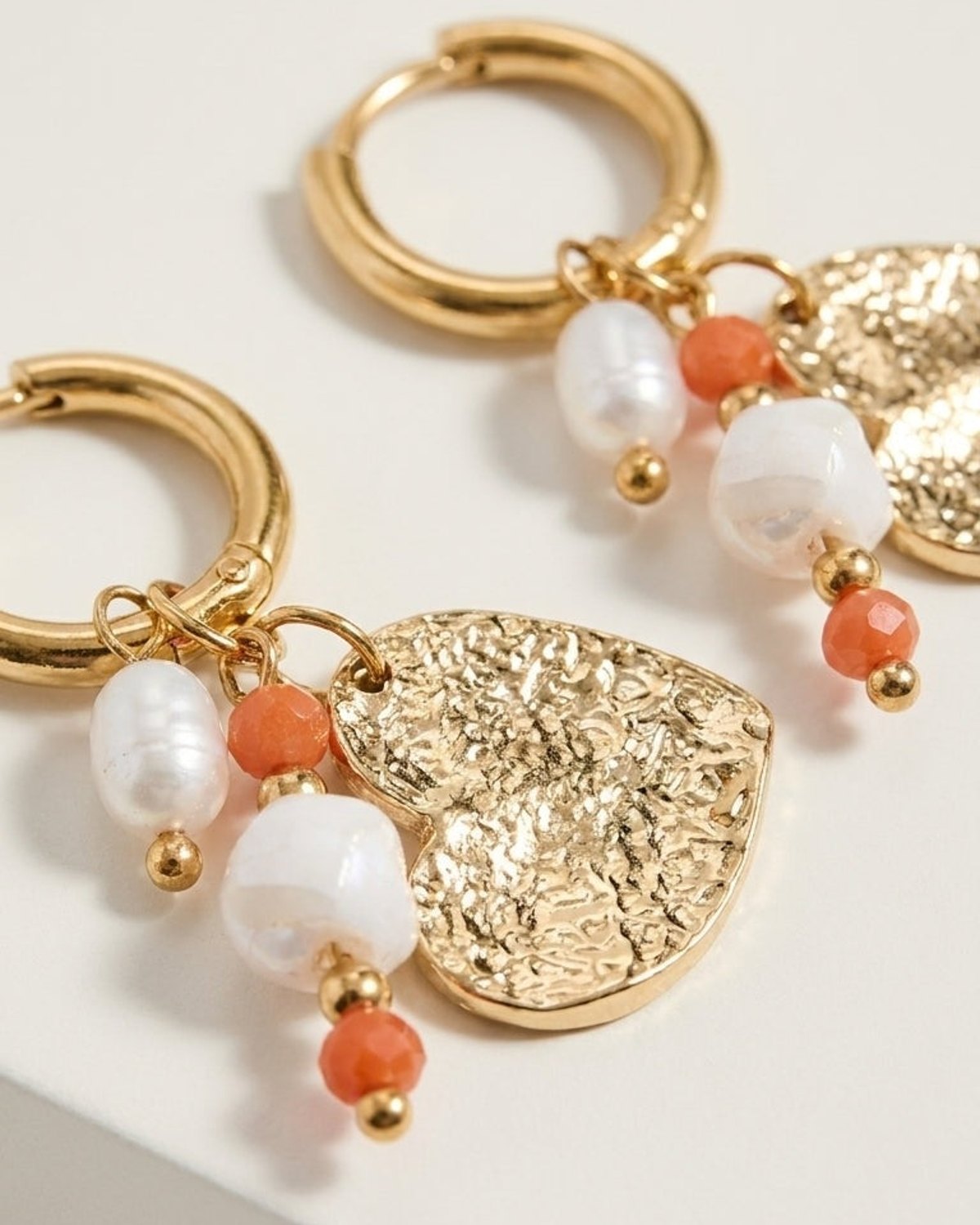 Gold hoop earrings featuring textured heart charms, white pearls, and orange beads on a white surface.