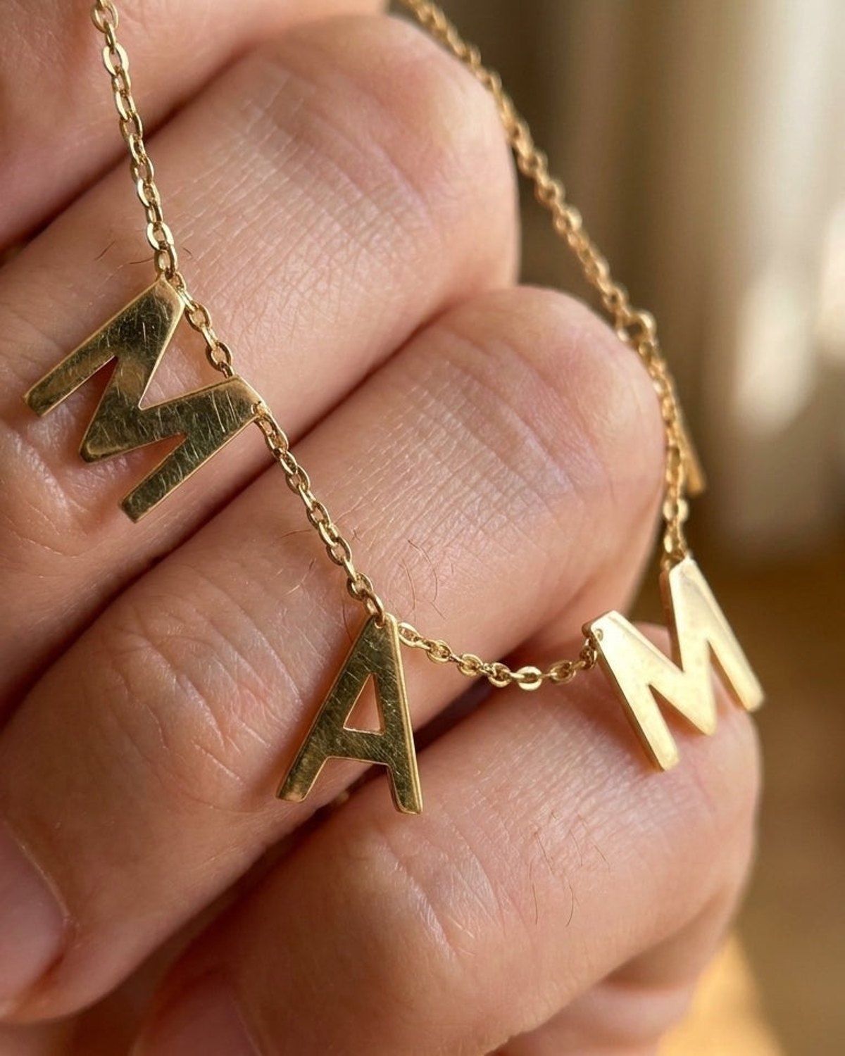 Gold chain necklace with brushed metal charms spelling "M A M" held in a hand.