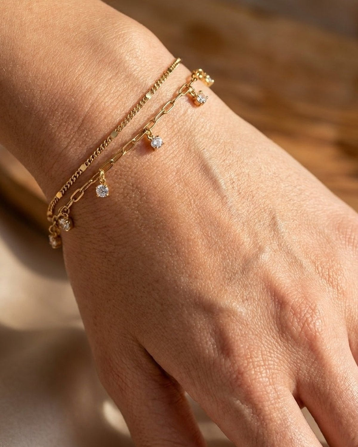 Two-layer gold chain bracelet with small hanging crystal charms worn on a wrist.