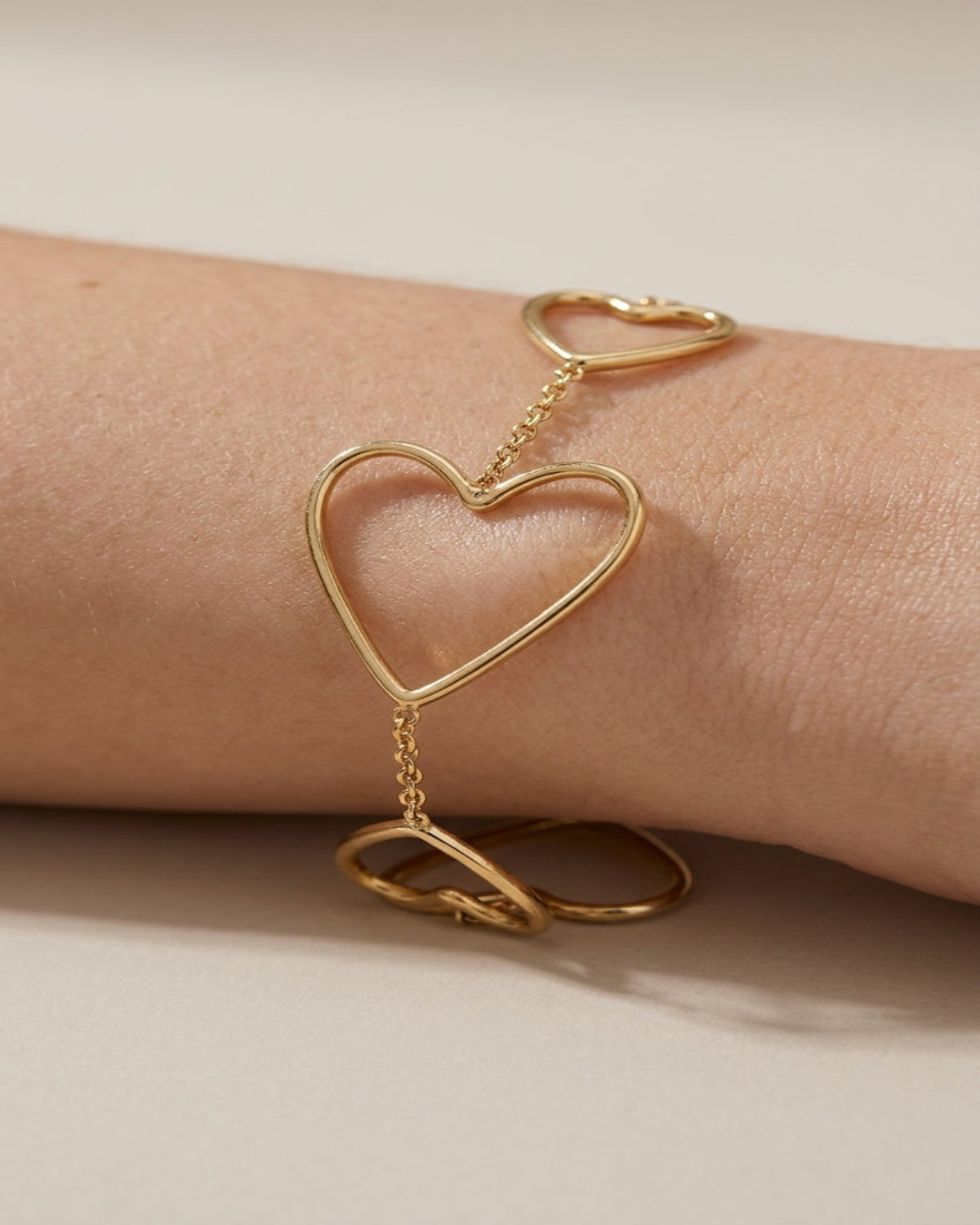 A gold heart-shaped open-link bracelet with a delicate chain and ring details, shown worn on a person's wrist against a warm beige background.