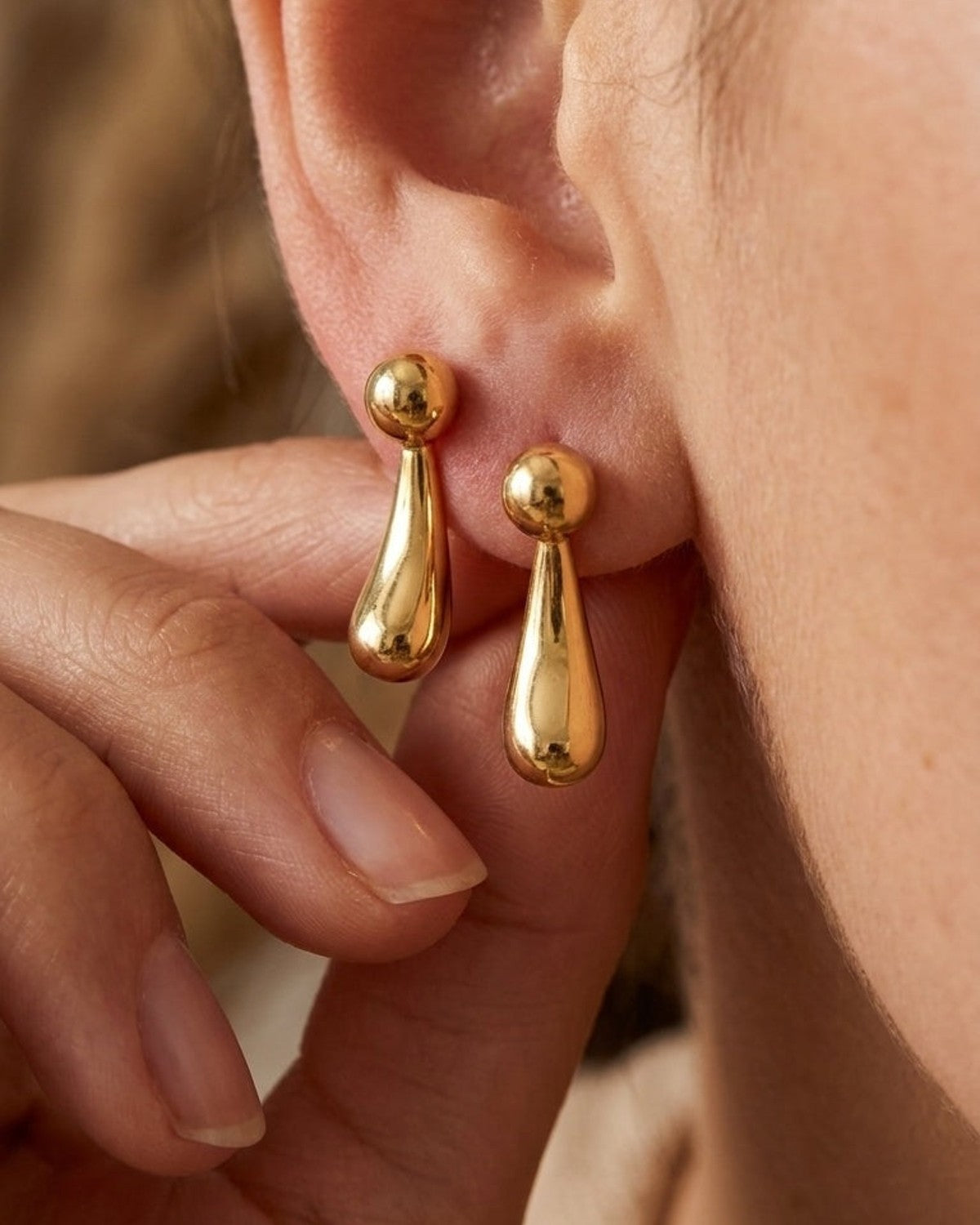 Polished gold drop earrings shaped like teardrops beneath small spheres, worn on an earlobe.