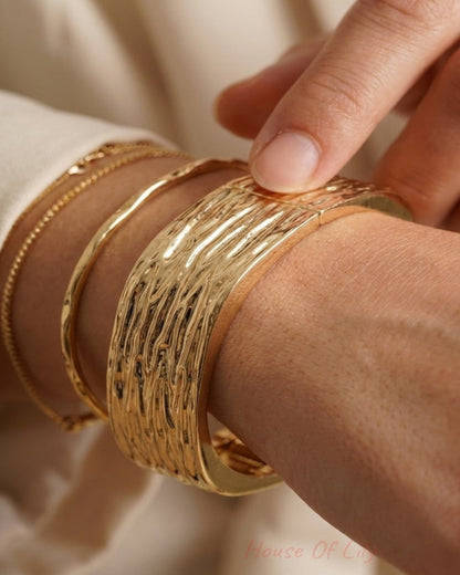 Close-up of a wide, textured gold cuff bracelet worn alongside thinner gold bangles on a wrist.