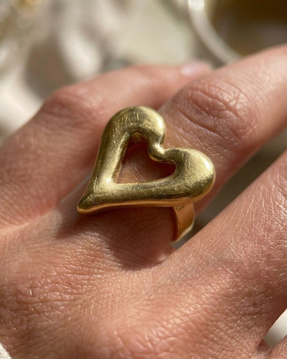 A gold-toned openwork heart ring worn on a hand.