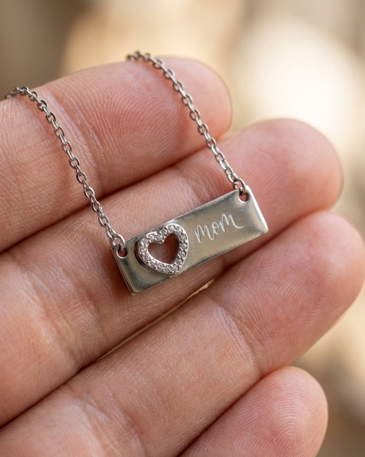 Silver chain necklace with a horizontal bar pendant inscribed with "Mom" and a pave heart cutout held in hand.