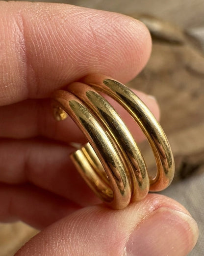 Fingers hold a pair of shiny, thick gold hoop earrings with a triple-band design.