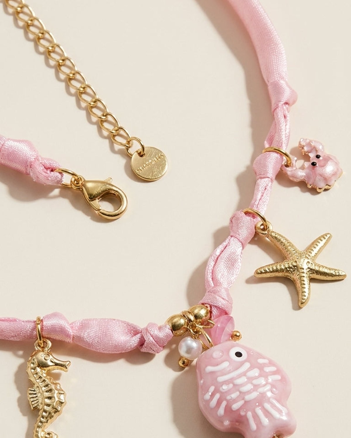 Pink braided ribbon necklace with gold-tone hardware, featuring ocean charms including a pink fish, seahorse, starfish, and crab.