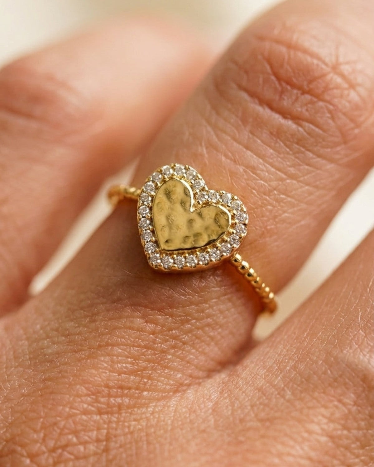 Gold ring with a hammered heart centerpiece framed by small sparkling zircon stones, worn on a finger.