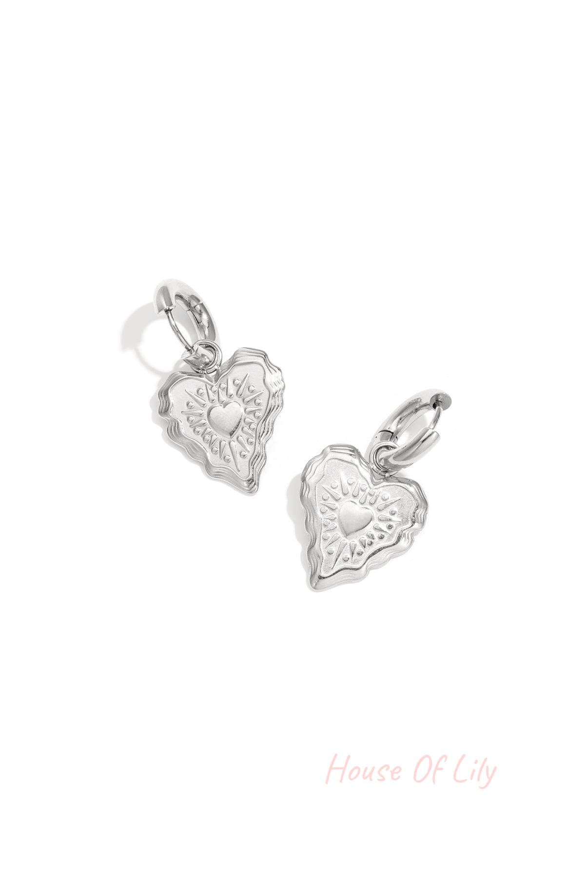 Close-up of silver heart charm hoop earrings with embossed detailing on a white background