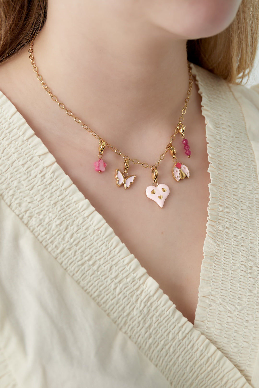 Close-up of woman wearing gold chain necklace with pink heart, butterfly, ladybug charms on soft cream blouse