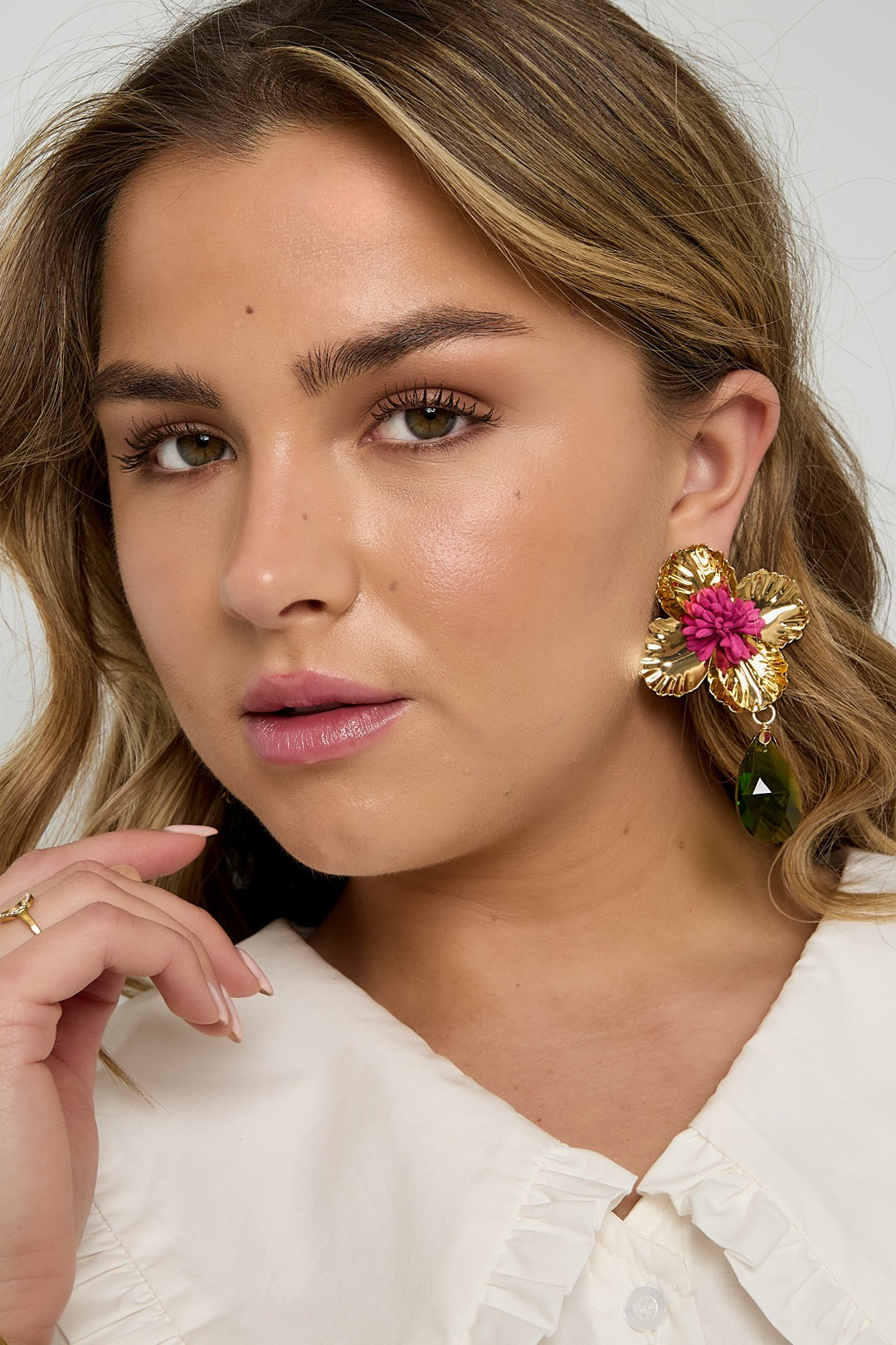 Close-up of woman wearing gold floral drop earrings with pink flower center and green teardrop pendant, hand near face