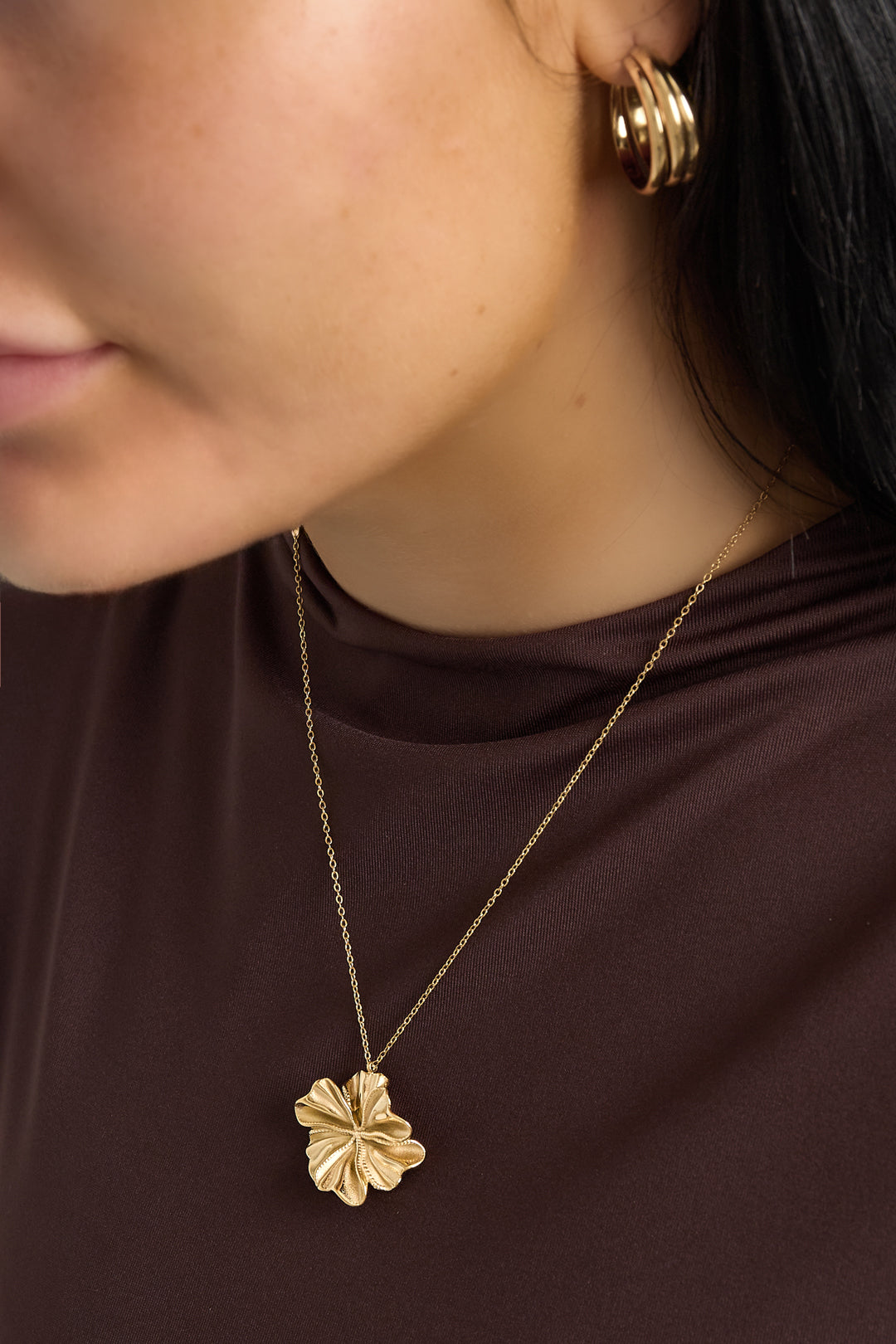 Close-up of woman wearing gold flower pendant necklace and matching gold hoop earrings with brown top