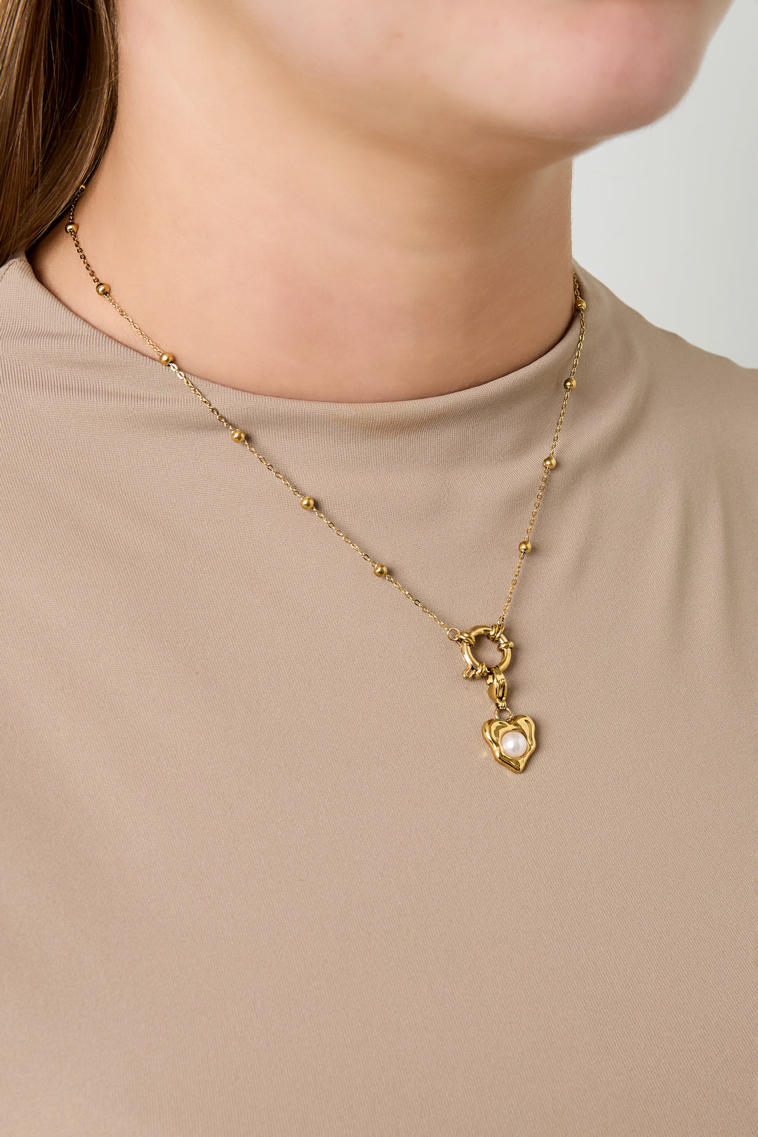 Woman wearing gold chain necklace with circular clasp and hanging heart charm featuring pearl detail, styled on beige top
