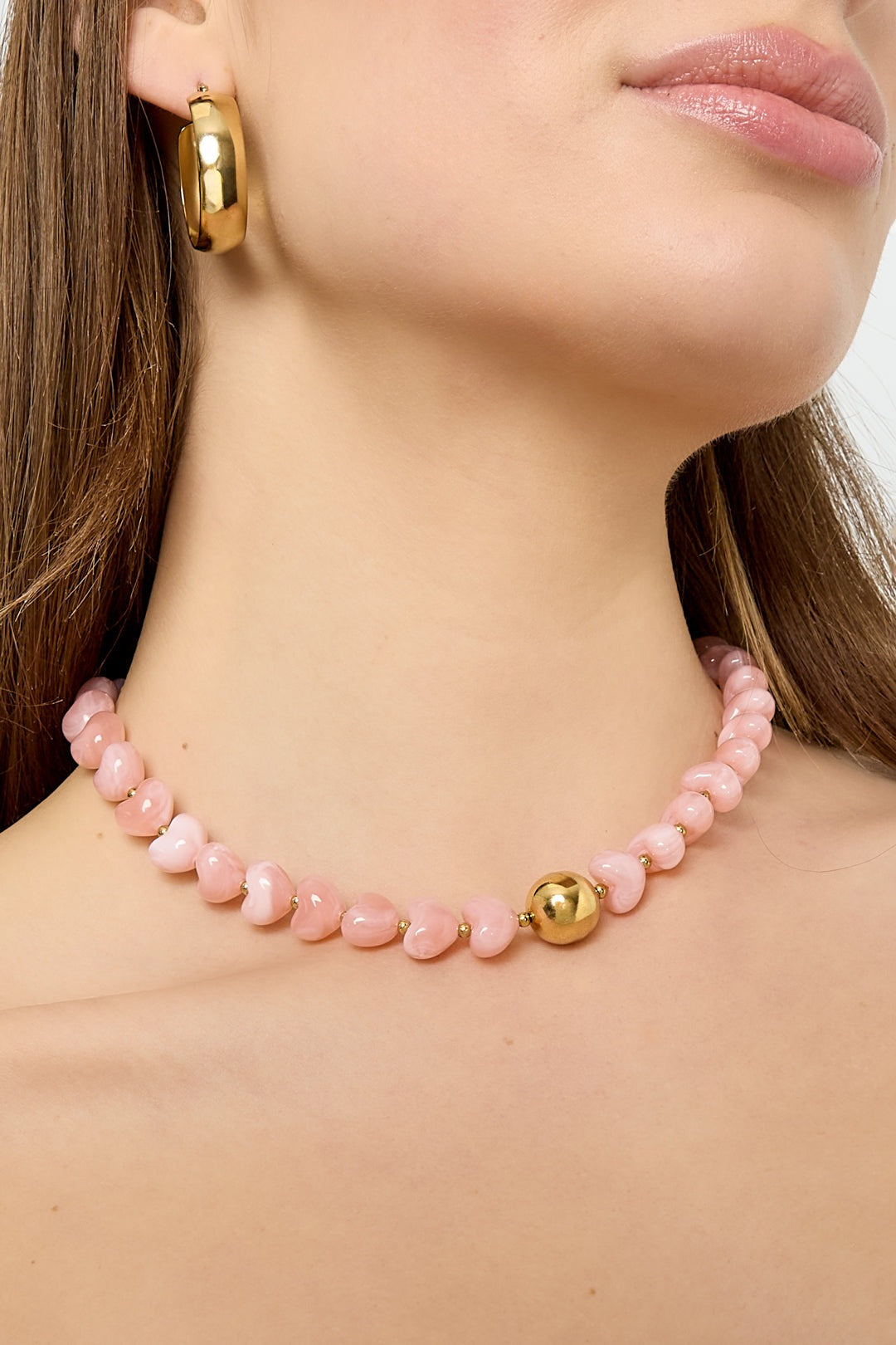 Side view close-up of model wearing the pink heart-shaped beaded necklace and gold hoop earrings showing the necklace’s glossy texture