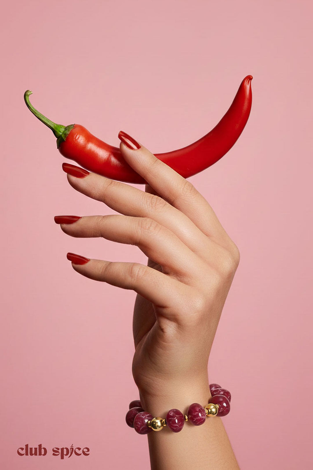 Woman's hand holding two red chili peppers wearing a vibrant red resin beaded bracelet with gold accents on a pink background